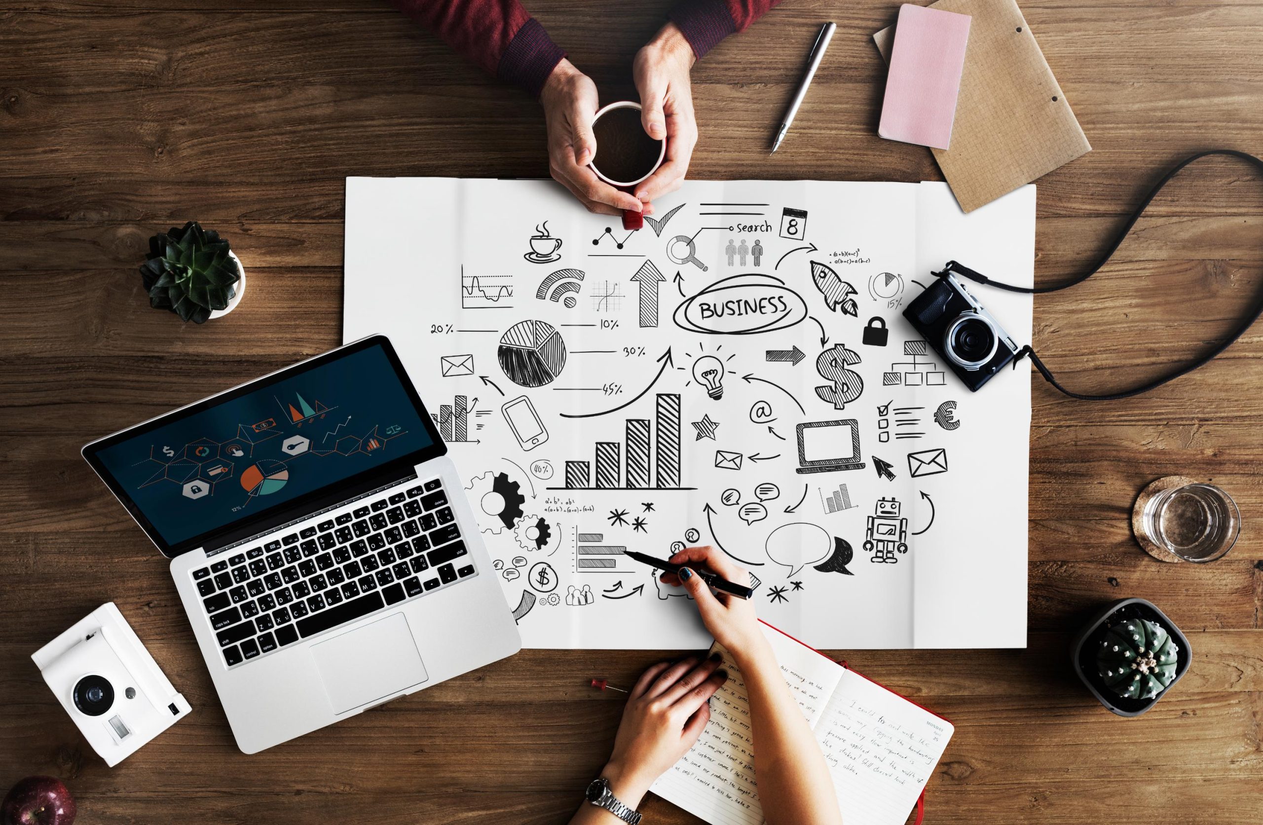 Creative business workspace with laptop displaying analytics, doodle-filled strategy sheet, notebook, camera, and hands collaborating at a wooden desk